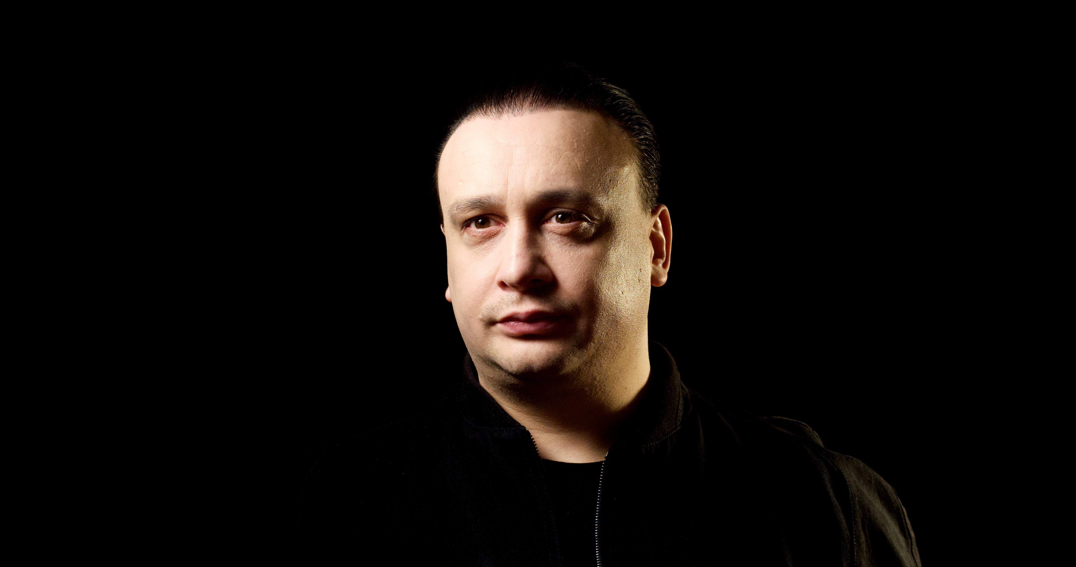 A studio portrait of Michael Darius Eastwood against a deep-black background, lit from the right, his head and shoulders squared to the camera and turned very slightly, wearing a black zip-front bomber jacket over a dark shirt, expression steady and neither smiling nor grim. Used as the hero backdrop for the Eden Protocol page.