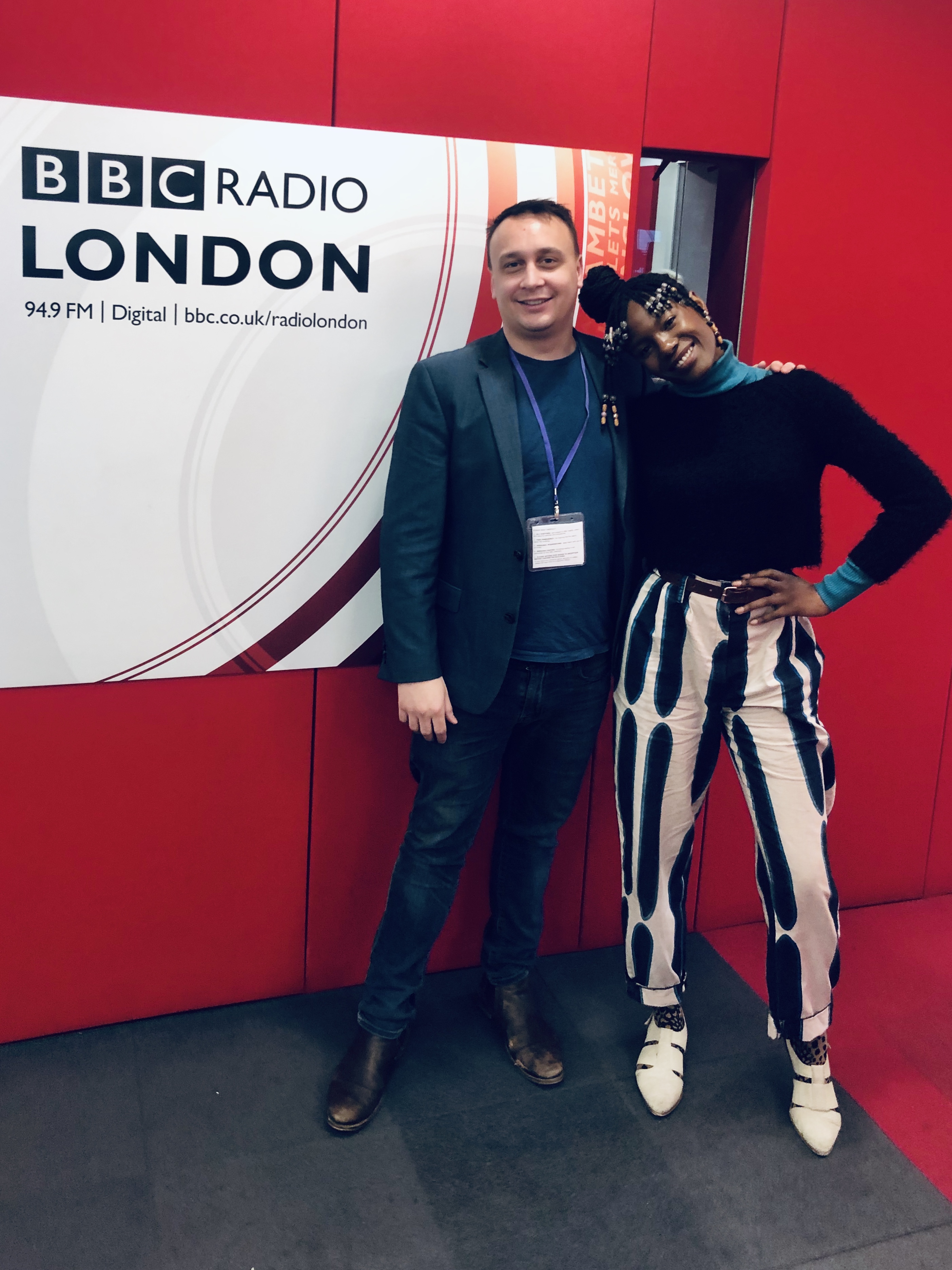 Michael Darius Eastwood wearing a visitor lanyard beside Shingai of the Noisettes in front of the BBC Radio London wall