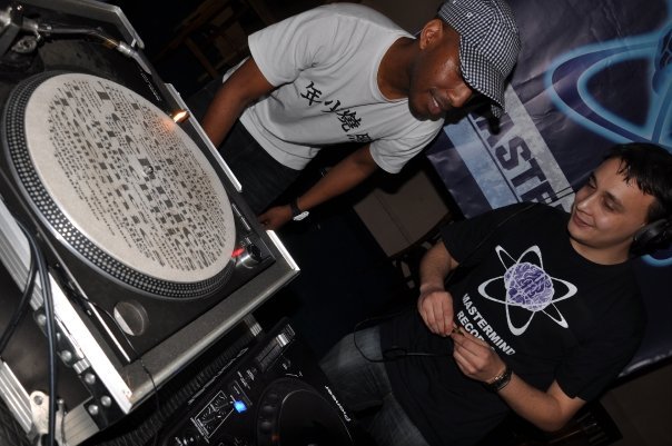 Michael Darius Eastwood at the turntables with Shy FX standing over the deck, a Mastermind Records banner behind and Michael wearing a Mastermind Records shirt